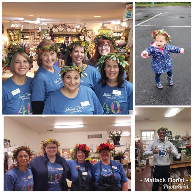 A collage of photos from a floral crown workshop. On the top left, six women in matching blue shirts smile as they stand in the store wearing floral crowns. On the top right, a toddler stands in the parking lot wearing a blue raincoat and a floral crown. On the bottom left, four women in matching blue shirts smile as they wear floral crowns. On the bottom right, a man in a gray sweatshirt smiles and gives two thumbs up as he wears a floral crown.