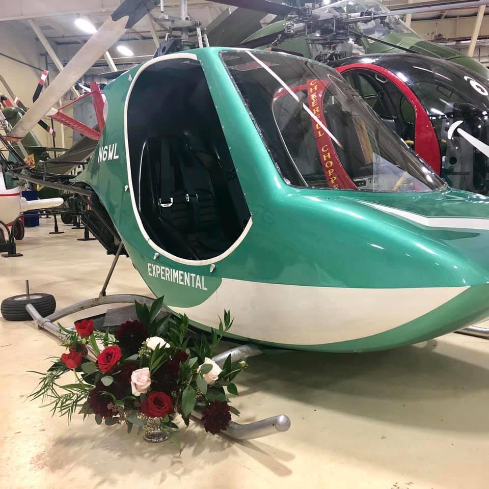 A small green and white helicopter with decals saying "EXPERIMENTAL" and "N6WL" sits in a warehouse with other helicopters. A white and red floral arrangement in a glass vase sits on the ground next to the helicopter.