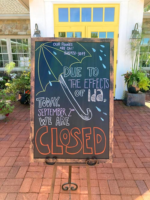 A handwritten chalkboard sign stands outside the front doors of the store that says "Due to the effects of Ida, today, September 2nd we are closed." The sign has a picture of an umbrella in the background.
