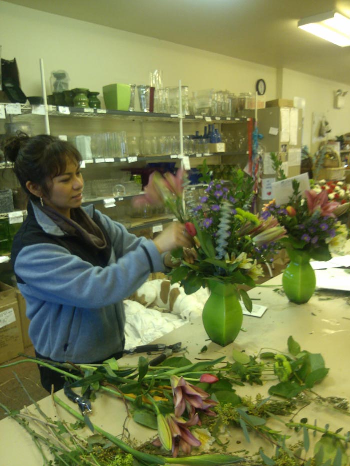 A designer adds flowers to an arrangement in a green vase. She is wearing a blue and black jacket and her hair is in a bun. On the table in front of her is a pile of flower stems and knives. On the wall behind her is a large shelf of vases.