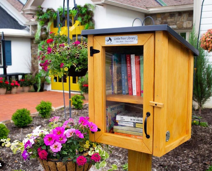 A small wooden box on a post sits in front of the storefront. The box is filled with books and has a plaque that reads "Little Free Library: take a book, share a book." The box is surrounded by hanging baskets of plants in a variety of colors.