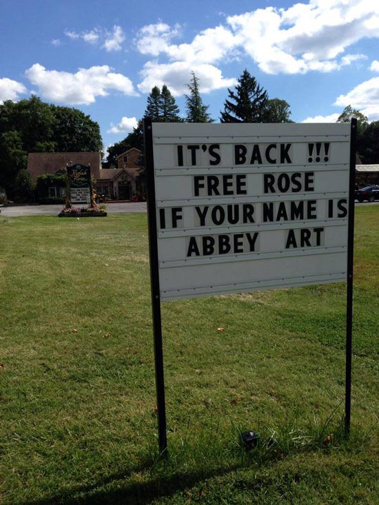 A sign sits in the grass outside that reads "It's back!!! Free rose if your name is Abbey Art"
