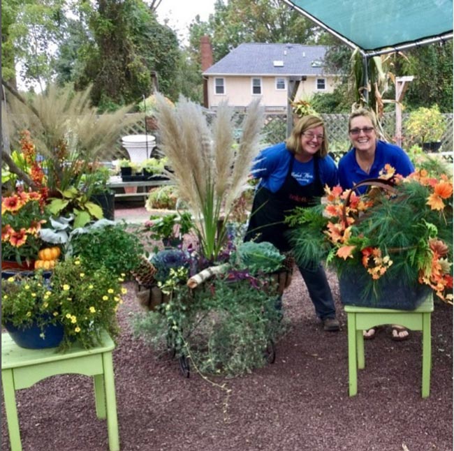 Two women in blue shirts stand behind a large outdoor plant and smile.