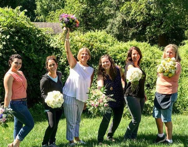 Six women stand in a grassy area. They are smiling and holding wedding bouquets.