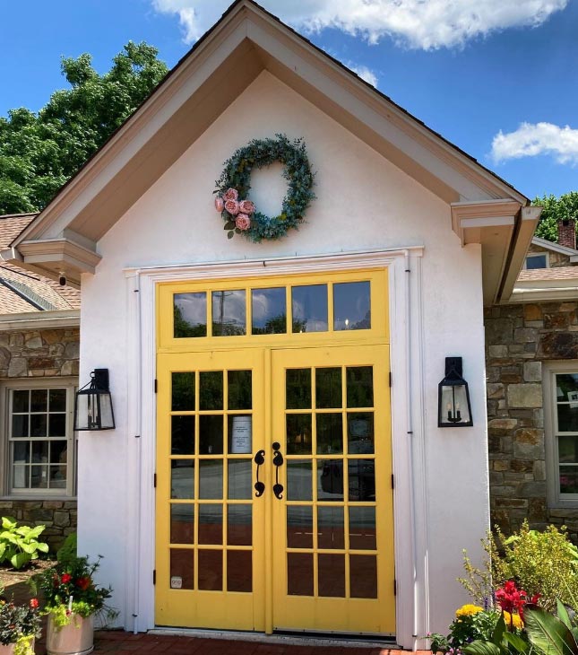An image of the double doors at the front of the store. The doors are painted yellow, and there is a green wreath with pink roses hanging above them.